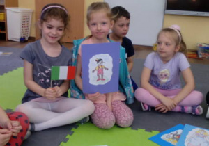 Dwie dziewczynki trzymające flagę Włoch i obrazek Pinokia, siedzące na dywanie wśród dzieci